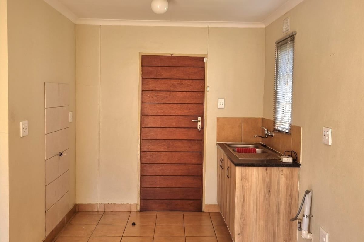 A compact, empty kitchen area featuring tiled flooring, built-in cabinetry with a stainless steel sink, and a wooden door leading to the exterior.