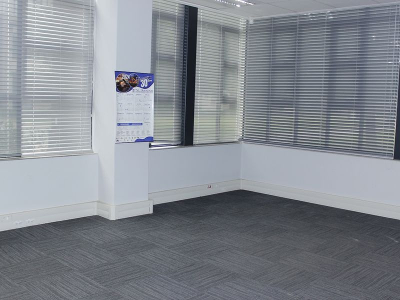 A bright, empty commercial office space featuring grey carpet tiles, large windows with venetian blinds, and a clean, professional finish.