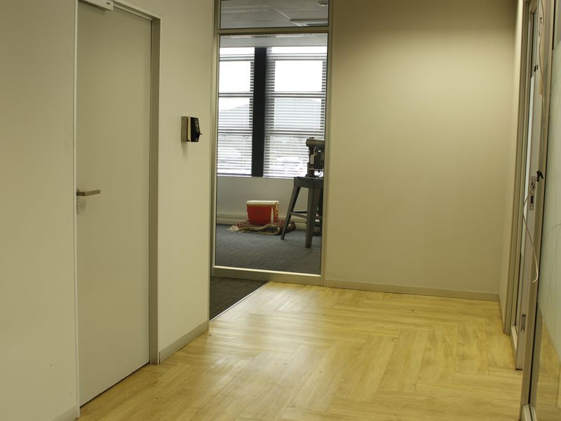 A clean, modern commercial hallway featuring herringbone vinyl flooring, glass partitioning, and secure access control, leading into an adjacent office space.