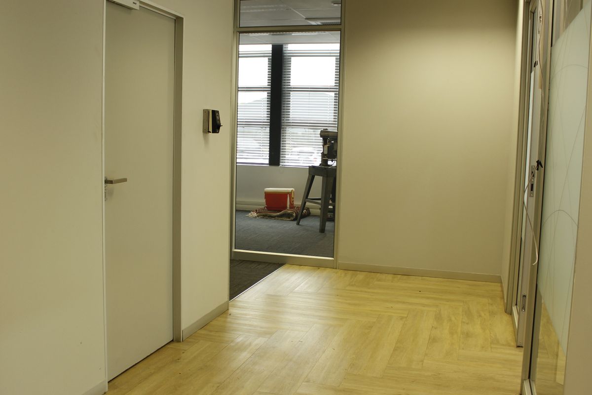 A clean, modern commercial hallway featuring herringbone vinyl flooring, glass partitioning, and secure access control, leading into an adjacent office space.