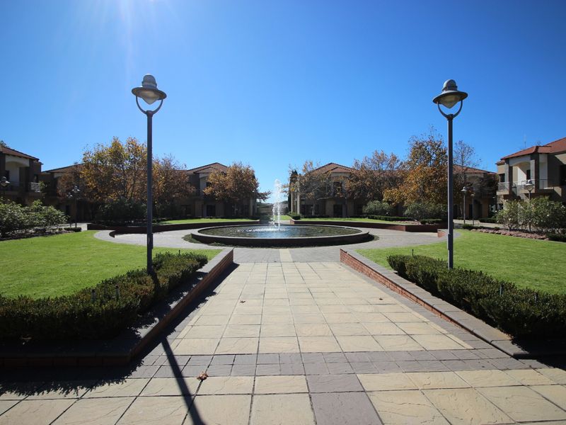 A well-maintained commercial courtyard featuring a central fountain, paved walkways, manicured lawns, and surrounding multi-story building facades.