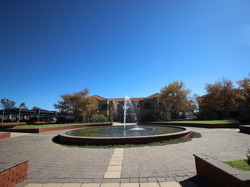 A well-maintained commercial office park entrance featuring a central circular water fountain, brick paved courtyard, and landscaped garden areas.