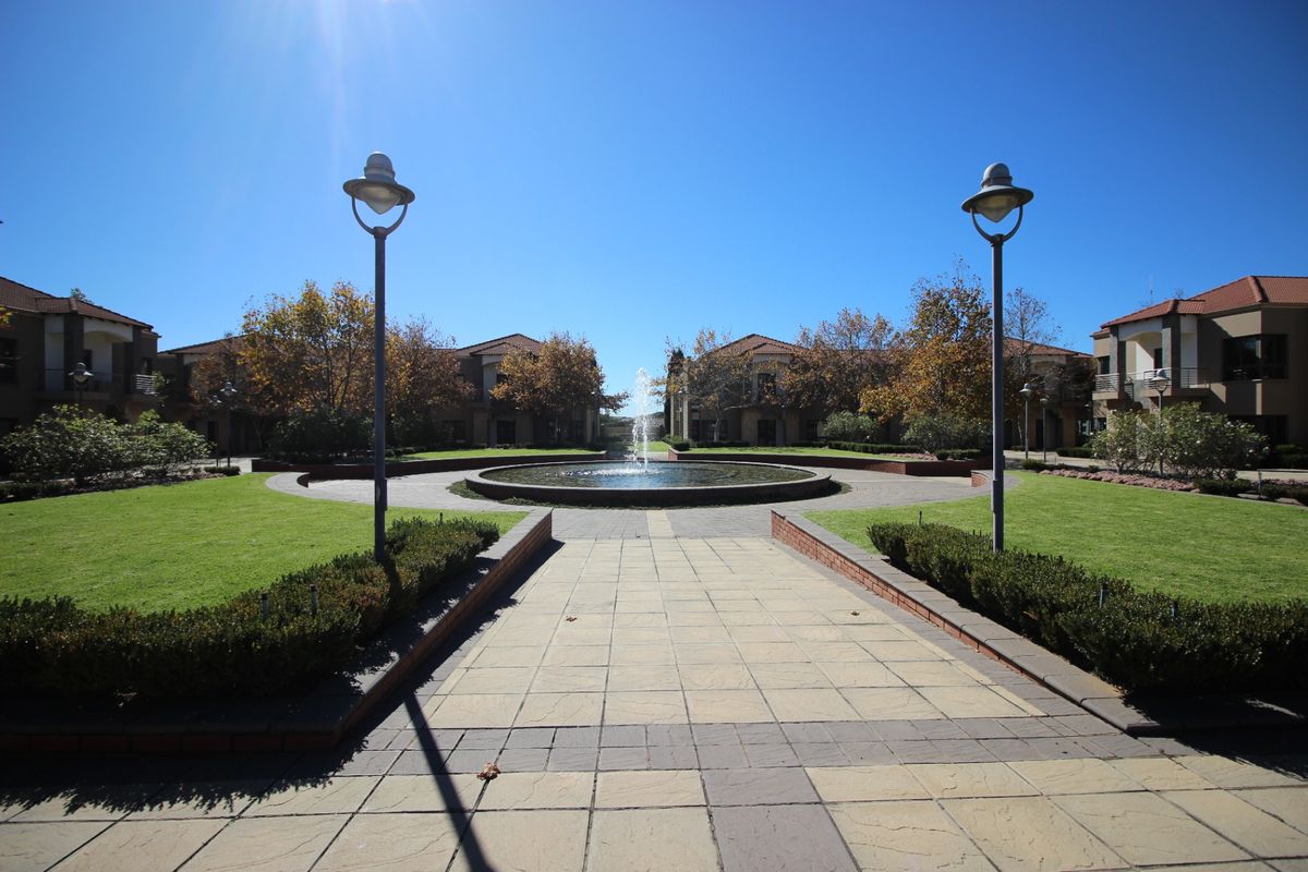 A well-maintained commercial courtyard featuring a central water fountain, paved walkways, manicured lawns, and surrounding office building facades.