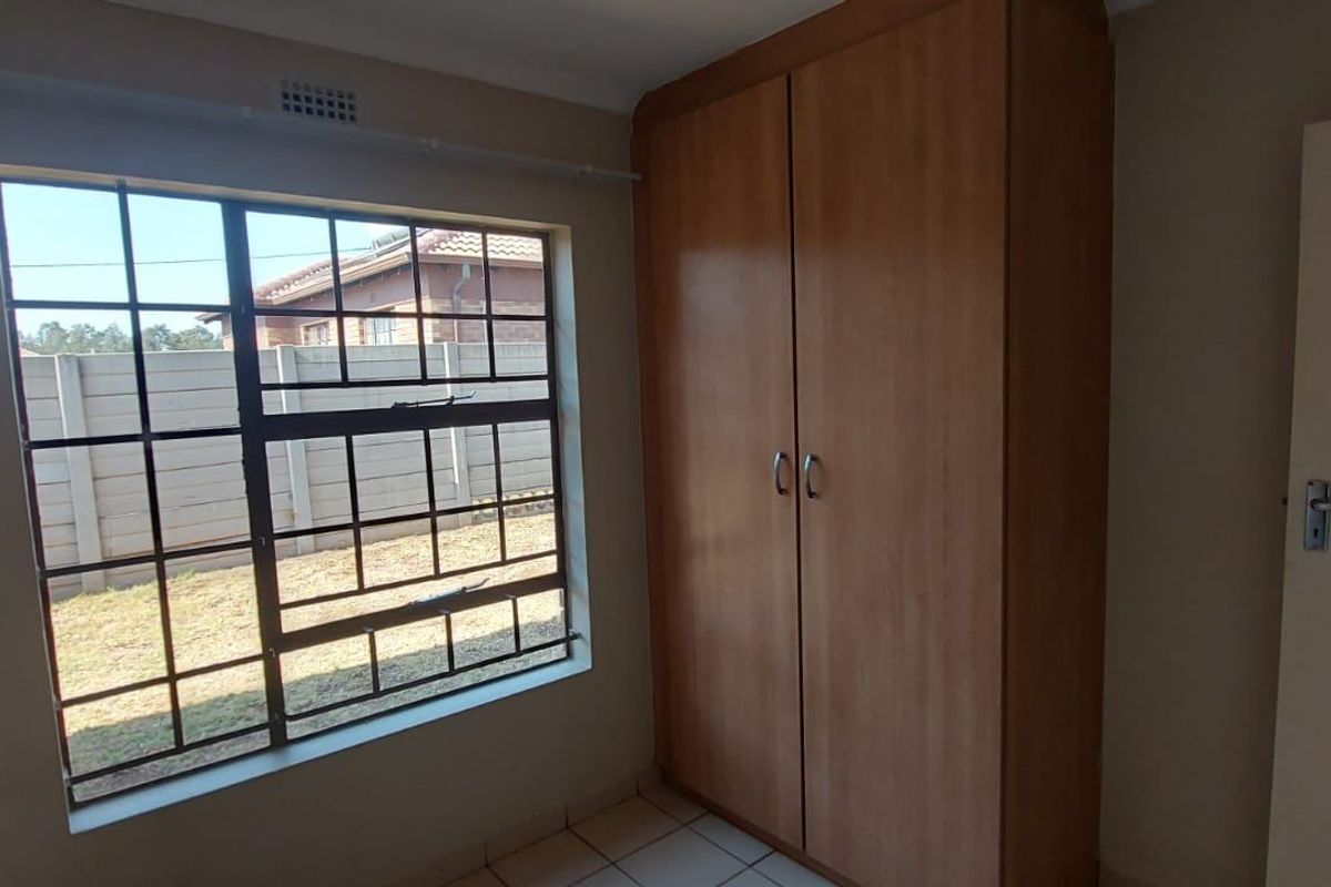 A clean, empty bedroom featuring tiled flooring, a built-in wooden wardrobe, and a window fitted with security bars.