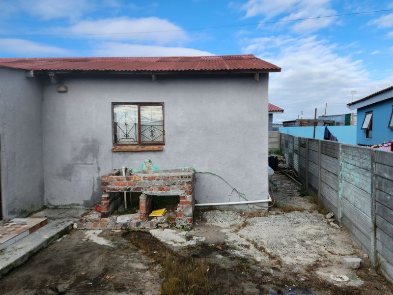 Rear exterior view showing a basic dwelling with a corrugated roof, precast boundary wall, and an unfinished brick outdoor wash basin area.