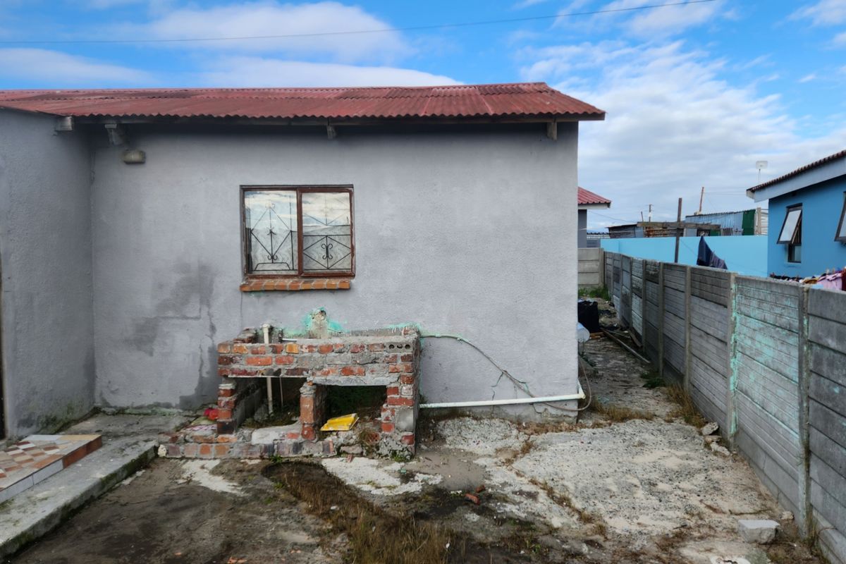 Rear exterior view showing a basic dwelling with a corrugated roof, precast boundary wall, and an unfinished brick outdoor wash basin area.