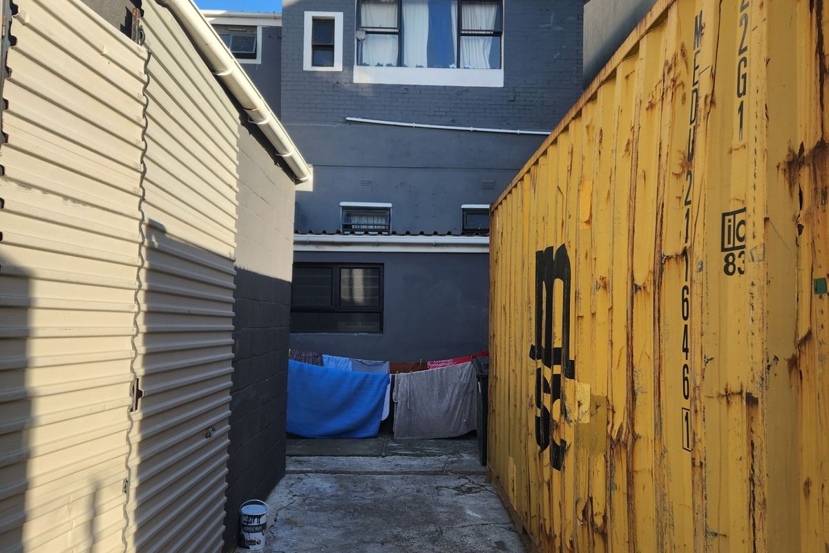 A narrow rear exterior passage between a corrugated metal structure and a shipping container, leading to the rear facade of the main building.