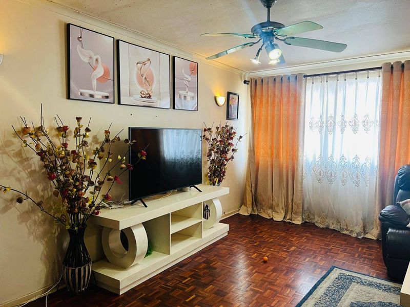 A cozy living room featuring classic parquet flooring, a ceiling fan, and large windows with curtains, providing a comfortable and functional living space.