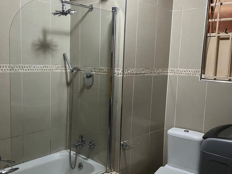 A functional bathroom featuring a tiled bathtub with a glass shower screen, a toilet, and exposed copper plumbing fixtures.