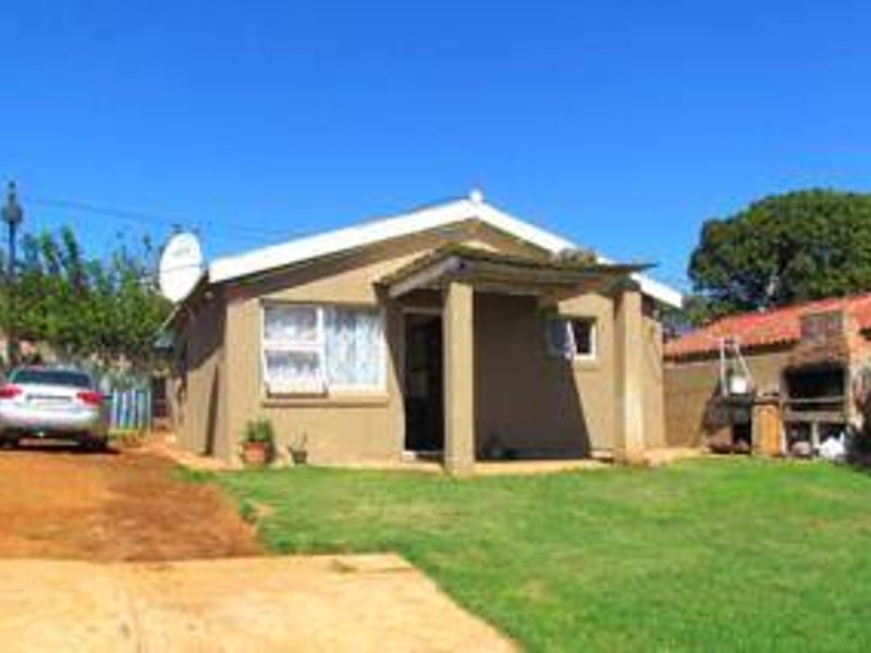 Front view of a single-story house featuring a covered porch, a brick braai area, and a lawn with an unpaved driveway.