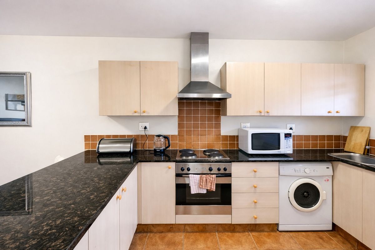 A functional kitchen featuring light wood cabinetry, dark granite countertops, a tiled backsplash, and integrated appliances including an oven, hob, and washing machine.