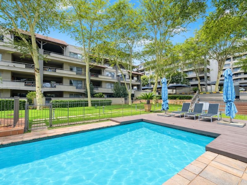 A well-maintained communal swimming pool area featuring timber decking, paved surrounds, and landscaped gardens within an apartment complex setting.