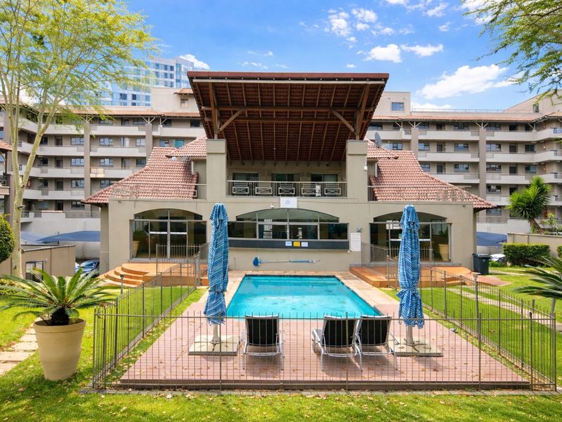 A well-maintained communal swimming pool area featuring a tiled deck, metal fencing, and a covered patio structure within an apartment complex.