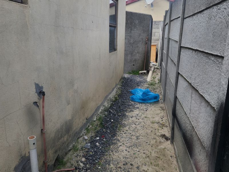 A narrow side passage of the house featuring a precast concrete boundary wall, gravel ground cover, and exposed exterior plumbing fixtures.