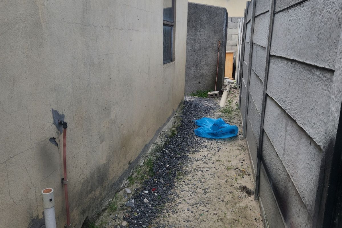 A narrow side passage of the house featuring a precast concrete boundary wall, gravel ground cover, and exposed exterior plumbing fixtures.