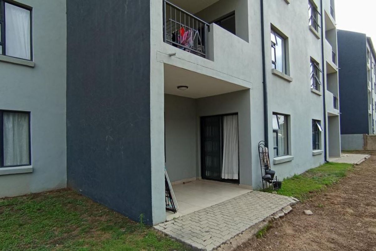 Rear view of the apartment building featuring a ground-floor tiled patio, sliding glass doors, and metal balcony railings with unfinished surrounding garden area.