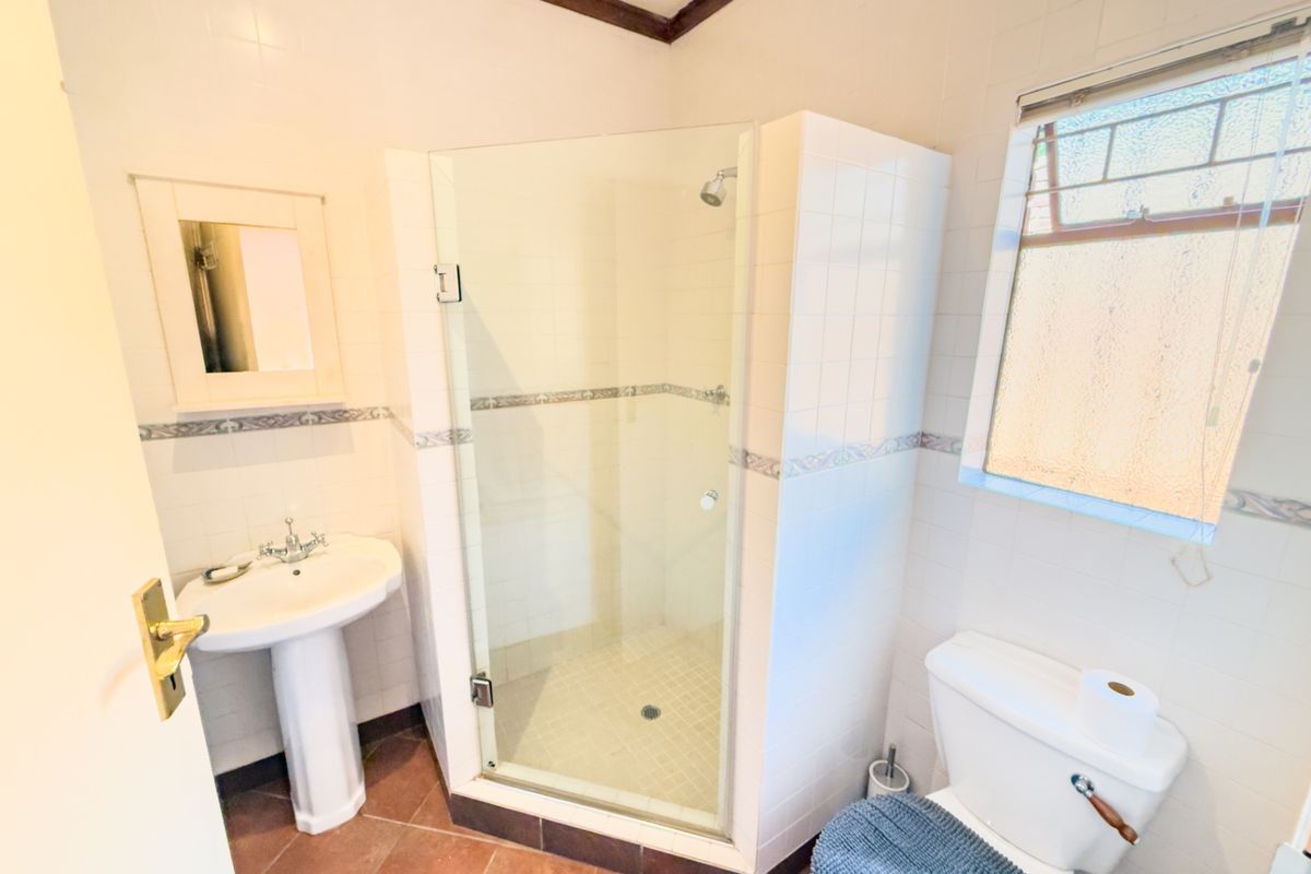 A functional bathroom featuring a pedestal basin, a glass-enclosed shower, and a toilet, finished with neutral wall tiles and terracotta-style floor tiles.