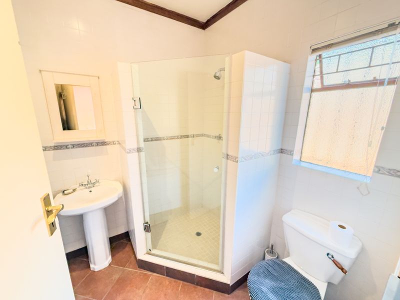 A functional bathroom featuring a pedestal basin, a glass-enclosed shower, and a toilet, finished with neutral wall tiles and terracotta-style floor tiles.