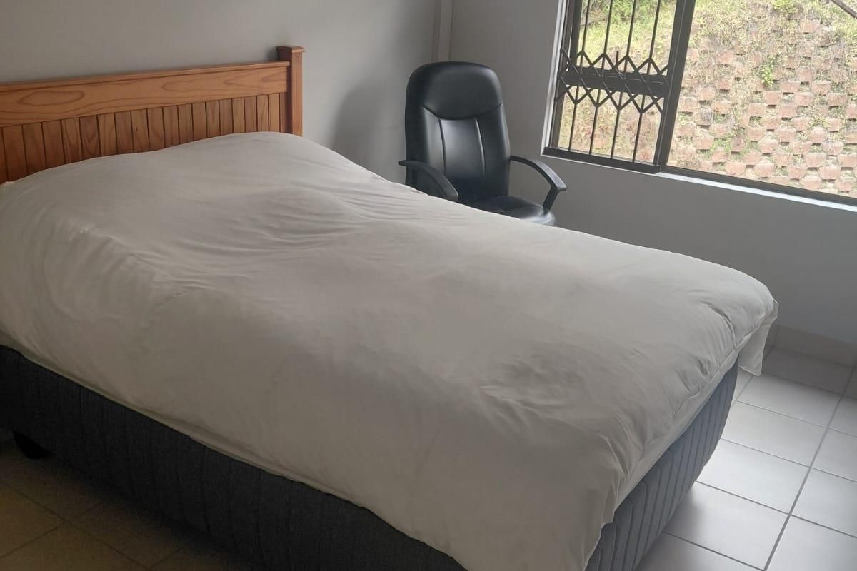 A functional bedroom featuring tiled flooring, a wall-mounted air conditioning unit, and a window fitted with a security gate and roller blind.