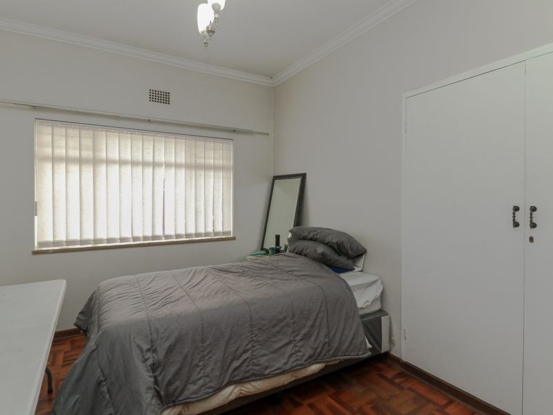 A bedroom featuring classic parquet flooring, a built-in wardrobe, and vertical blinds, offering a functional space with neutral wall finishes.
