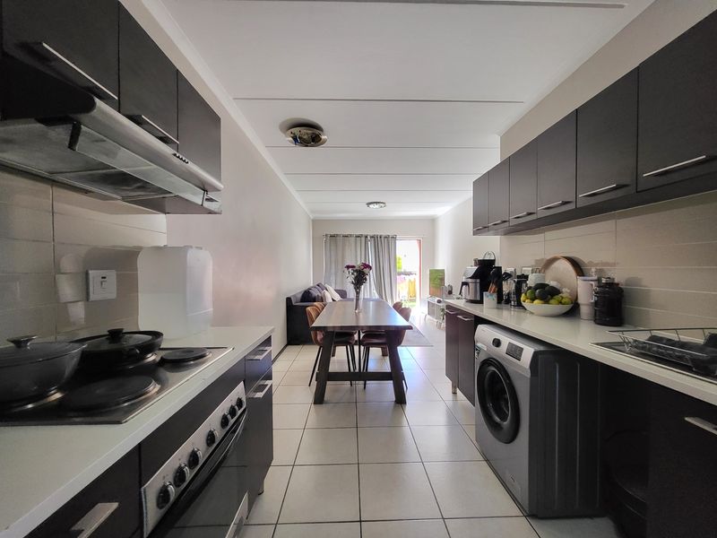 Open-plan kitchen and dining area featuring dark cabinetry, tiled flooring, and integrated laundry facilities, leading through to a living space with balcony access.