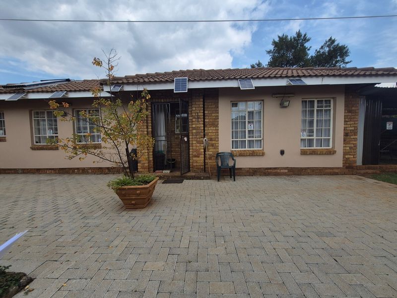 Front exterior view of a single-story brick house featuring a paved driveway, security gates, and roof-mounted solar panels.