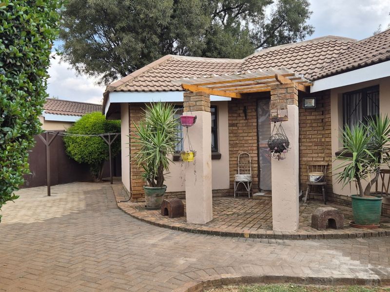 A rear exterior view of a brick house featuring a paved patio area with a pergola and established garden surroundings.