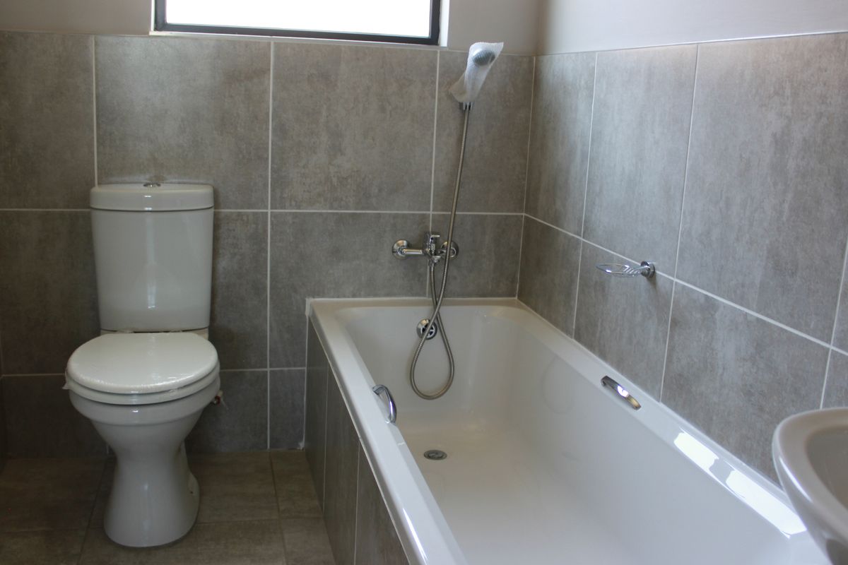 A clean, modern bathroom featuring neutral grey wall tiles, a white bathtub with a handheld shower mixer, and a standard toilet.