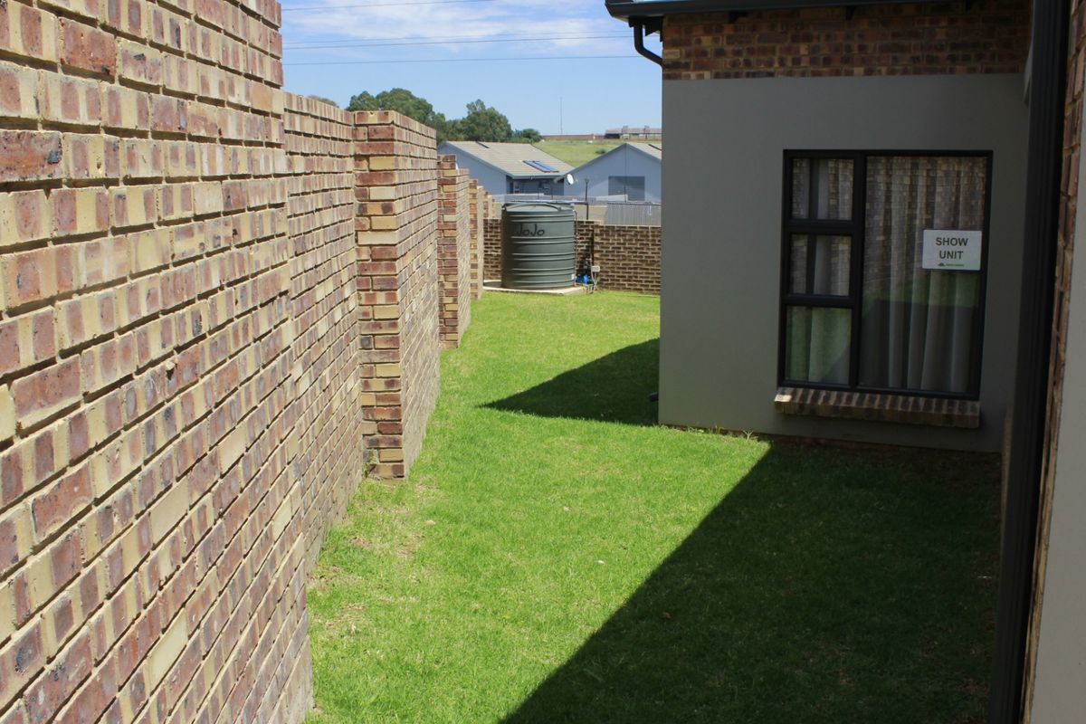 A side garden area featuring a face brick boundary wall, a water tank, and a modern plastered exterior wall with aluminium window frames.