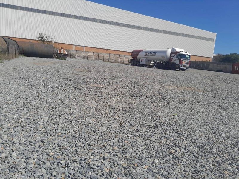 A large industrial gravel yard featuring perimeter fencing, storage tanks, and a view of the adjacent warehouse building exterior.