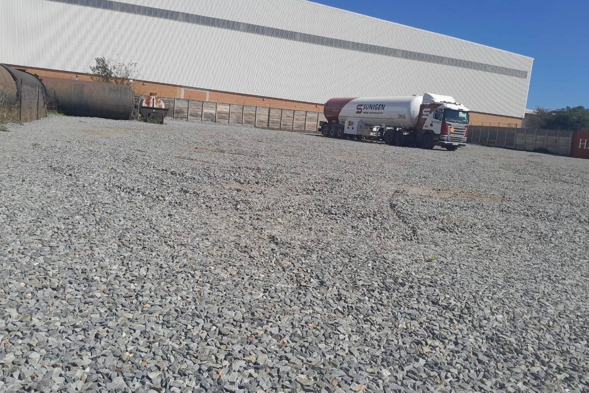 A large industrial gravel yard featuring perimeter fencing, storage tanks, and a view of the adjacent warehouse building exterior.