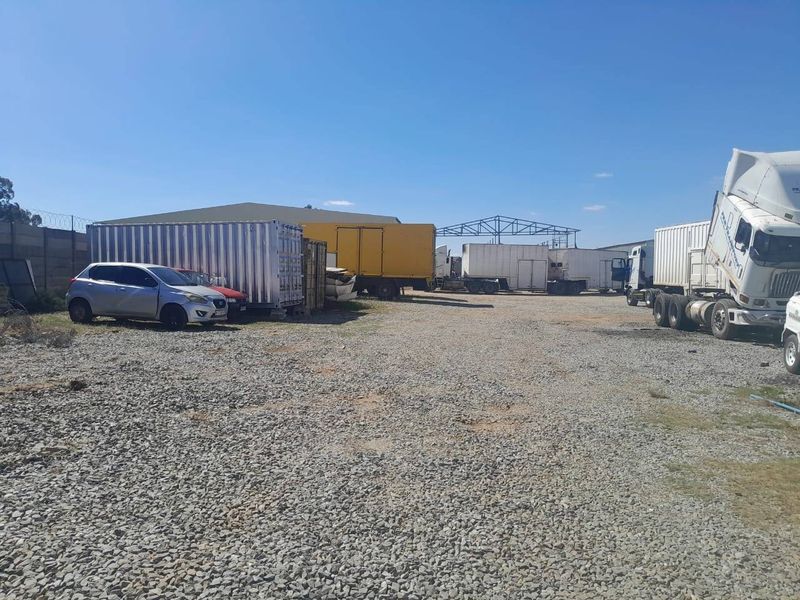 An expansive, unpaved commercial yard featuring a gravel surface, perimeter fencing, and an open-air steel structure suitable for industrial storage or logistics operations.