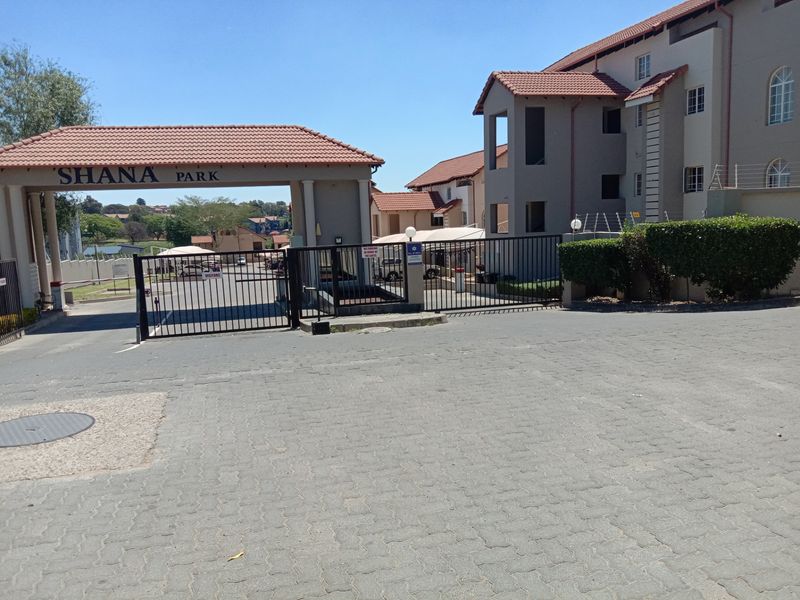 Entrance view of Shana Park apartment complex featuring a secure gated access point, paved driveway, and multi-story residential buildings with tiled roofing.