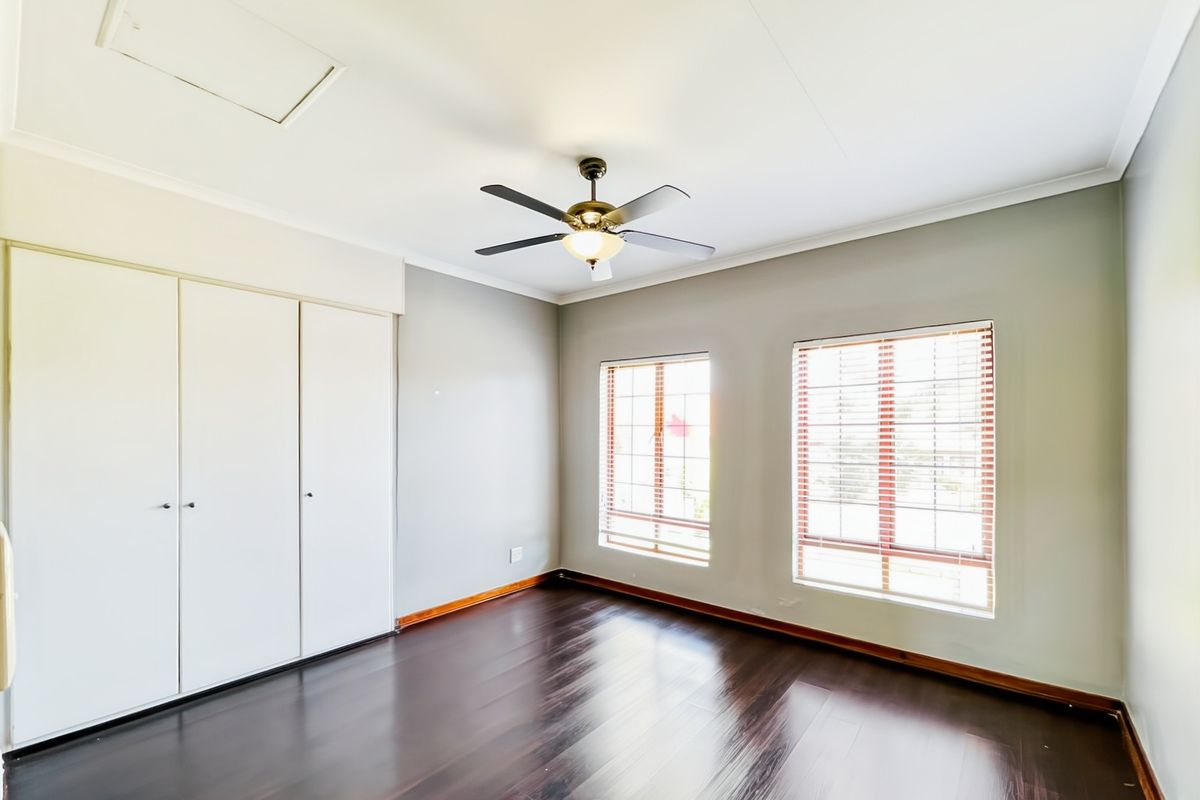 A bright, empty bedroom featuring dark wood-look flooring, built-in wardrobes, a ceiling fan, and large windows with fitted blinds.