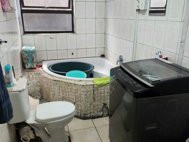 A compact bathroom featuring a corner bathtub, toilet, and integrated laundry space with tiled finishes throughout.