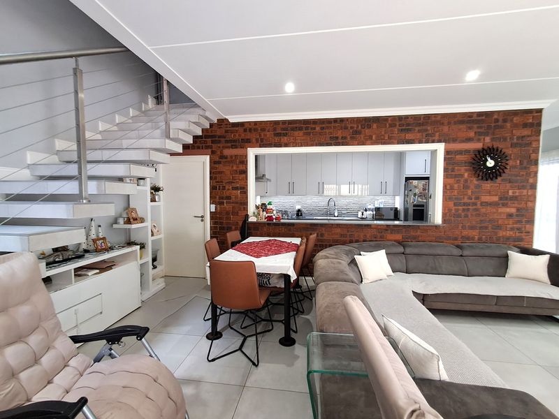 Open-plan living and dining area featuring a modern staircase, exposed brick feature wall, and a pass-through window connecting to the kitchen.