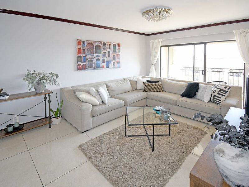 A bright and well-maintained living room featuring large format floor tiles, crown molding, and a sliding glass door leading to a balcony.