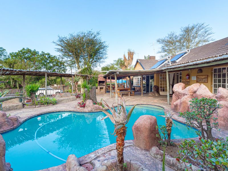 Outdoor pool area featuring a rock-scaped swimming pool, covered patio with built-in braai, and solar water heating system on the roof.