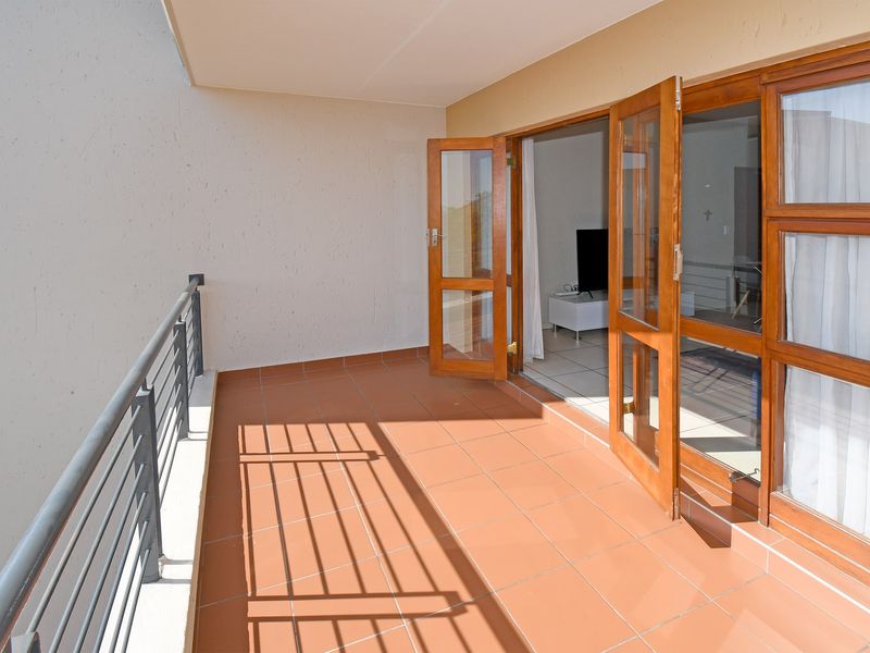 A spacious covered balcony featuring terracotta-style floor tiles, a metal safety balustrade, and wooden-framed glass doors leading into the interior living area.