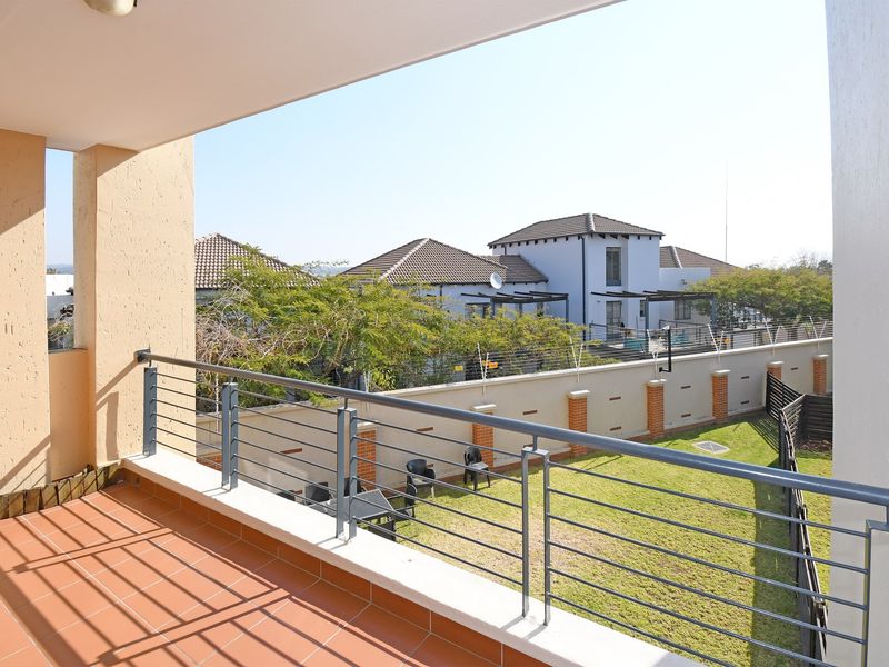 A spacious covered balcony featuring tiled flooring and a secure metal balustrade, overlooking a private garden area.