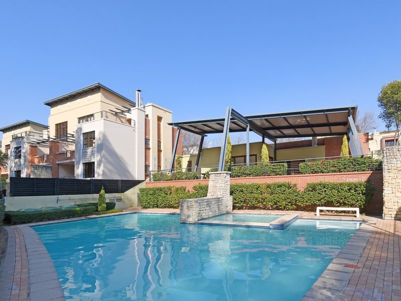 A well-maintained communal swimming pool area featuring a stone water feature, brick retaining walls, and a modern metal pergola for shaded outdoor relaxation.
