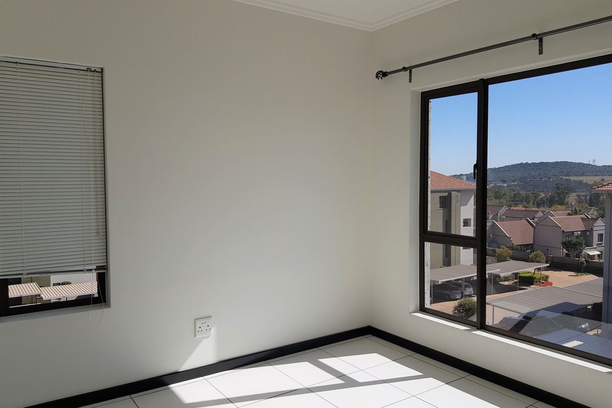 A bright, empty bedroom featuring white tiled floors, neutral walls, and large windows with views of the surrounding residential complex.