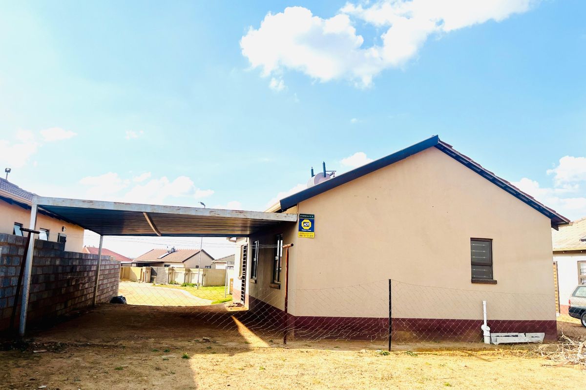 Rear view of the property featuring a metal carport structure, perimeter wall, and security fencing on an unpaved yard.