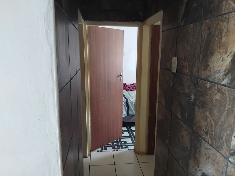 A narrow hallway featuring dark decorative wall tiles and light floor tiles, leading into an adjacent bedroom.