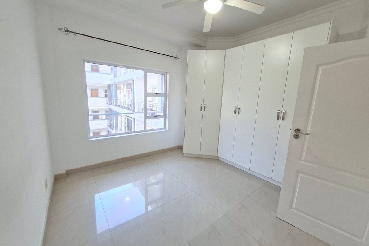 A bright, empty bedroom featuring neutral tiled flooring, built-in white cupboards, and a ceiling fan, offering a clean and versatile living space.