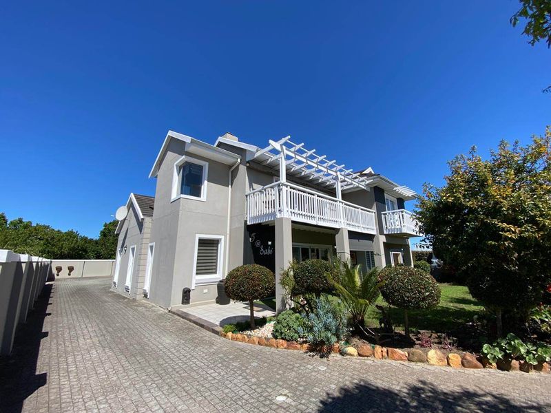 A well-maintained double-story residential property featuring a paved driveway, a prominent upper-level balcony with a timber pergola, and landscaped garden beds.