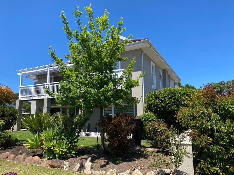A well-maintained two-story house exterior featuring a front balcony with a pergola, surrounded by established garden landscaping and stone garden edging.