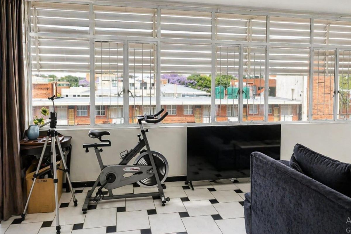 A sunlit living area featuring large windows with security bars, tiled flooring, and an urban neighborhood view.