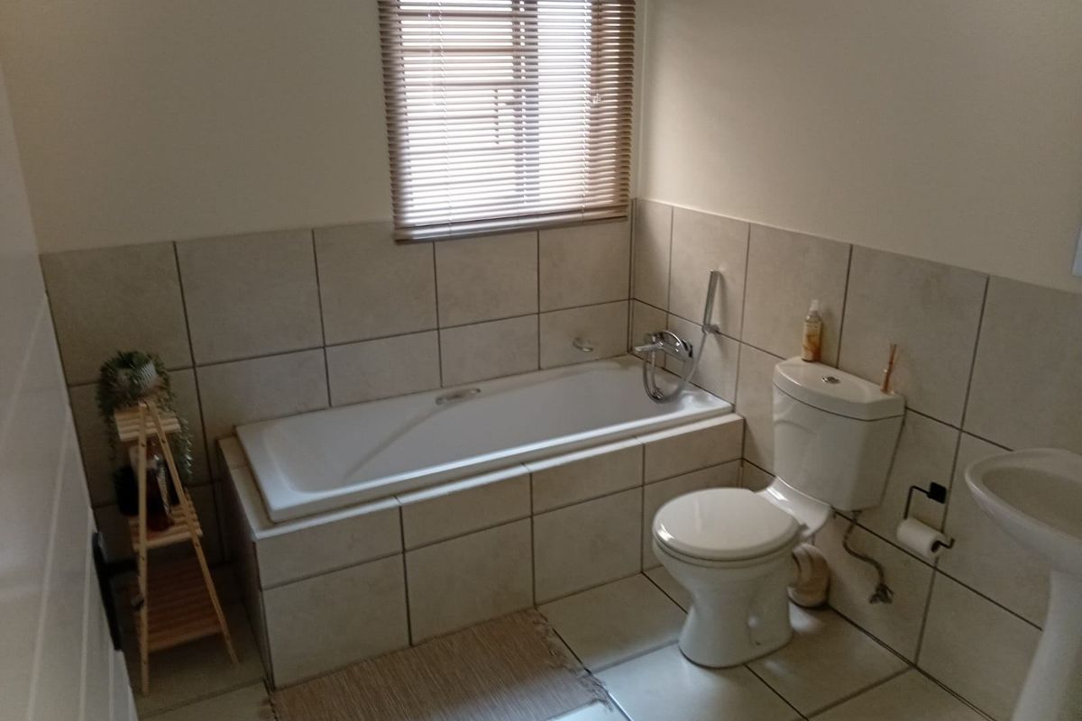 A functional bathroom featuring a built-in bathtub, pedestal basin, toilet, and neutral ceramic wall and floor tiling with a venetian blind.