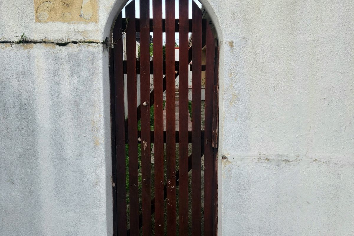 A weathered timber gate set within a boundary wall featuring visible plaster cracking and paint deterioration, topped with security barbed wire.